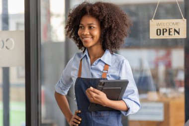 Successful african woman in apron standing coffee shop door. Happy small business owner holding tablet and working. Smiling portrait of SME entrepreneur seller business standing with copy space.