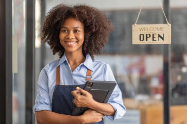 Successful african woman in apron standing coffee shop door. Happy small business owner holding tablet and working. Smiling portrait of SME entrepreneur seller business standing with copy space.