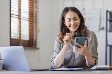 Photo of a joyful nice Asian indian woman using a smartphone with a laptop and smiling while sitting in workplace an home office.