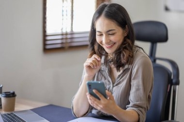 Photo of a joyful nice Asian indian woman using a smartphone with a laptop and smiling while sitting in workplace an home office.