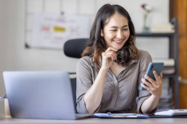 Photo of a joyful nice Asian indian woman using a smartphone with a laptop and smiling while sitting in workplace an home office.