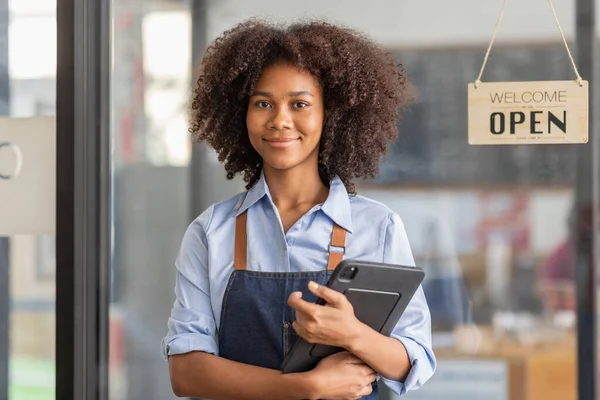 Successful african woman in apron standing coffee shop door. Happy small business owner holding tablet and working. Smiling portrait of SME entrepreneur seller business standing with copy space.
