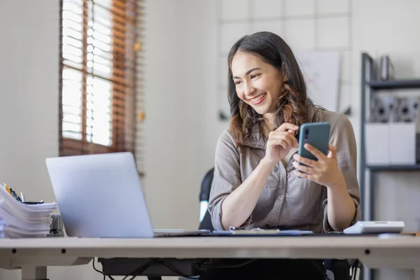Photo of a joyful nice Asian indian woman using a smartphone with a laptop and smiling while sitting in workplace an home office.