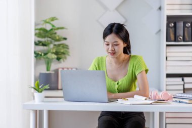 Çevrimiçi Ders. Gülümseyen Asyalı kadın dizüstü bilgisayar kullanıyor ve masasında otururken not alıyor, gülümseyen Asyalı kadın uzaktan eğitimin ve uzaklığın tadını çıkarıyor, boş alan arıyor.