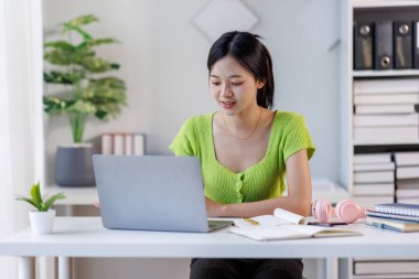 Çevrimiçi Ders. Gülümseyen Asyalı kadın dizüstü bilgisayar kullanıyor ve masasında otururken not alıyor, gülümseyen Asyalı kadın uzaktan eğitimin ve uzaklığın tadını çıkarıyor, boş alan arıyor.