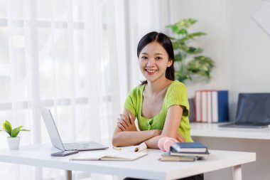 Çevrimiçi Ders. Gülümseyen Asyalı kadın dizüstü bilgisayar kullanıyor ve masasında otururken not alıyor, gülümseyen Asyalı kadın uzaktan eğitimin ve uzaklığın tadını çıkarıyor, boş alan arıyor.