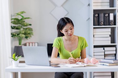 Çevrimiçi Ders. Gülümseyen Asyalı kadın dizüstü bilgisayar kullanıyor ve masasında otururken not alıyor, gülümseyen Asyalı kadın uzaktan eğitimin ve uzaklığın tadını çıkarıyor, boş alan arıyor.