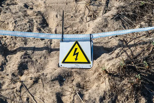 A yellow triangular warning sign for electrical hazard on sandy ground.
