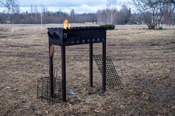 Bir barbekü ızgarası, ağaçlar, yeşil çimenler ve berrak mavi gökyüzünün altında tüylü beyaz bulutlarla çevrili yemyeşil bir tarlanın ortasına yerleştirilir.