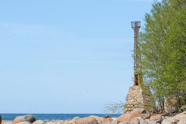 An old lighthouse tower stands beside the ocean shore, surrounded by rocks and greenery, under a clear blue sky.