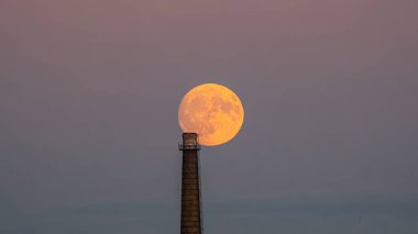 Dramatic shot of a full moon rising directly behind an industrial chimney against a twilight sky, blending natural and man-made elements. clipart