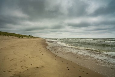 Kıyı şeridi boyunca uzanan terk edilmiş kumlu bir sahil dalgalar çarpıyor ve tepemizde kara bulutlar belirerek kasvetli bir atmosfer yaratıyor..