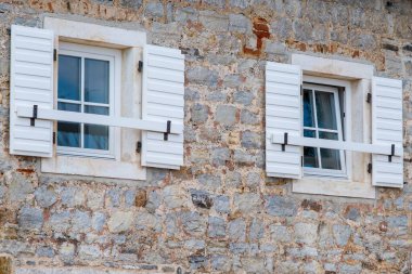Kırsal taştan bir duvarda iki beyaz pencere, geleneksel mimari elementleri büyüleyici ve zamansız bir tasarımla harmanlıyor..