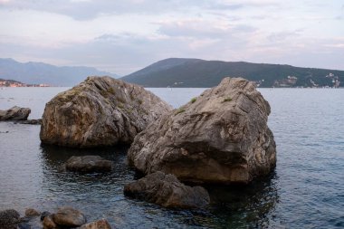 Alacakaranlıkta sakin sularda iki büyük kaya dinleniyor, uzak tepeler ve sakin bir kıyı arkaplanı var..