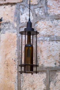 Metal bir çerçeveye sarılı, eşsiz bir şişe şeklindeki fener, desenli taş bir duvara asılı, kırsal ve klasik bir estetik sunuyor..