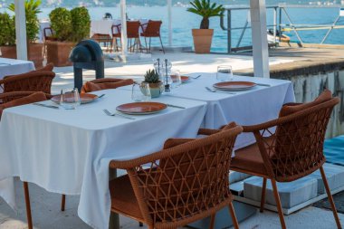 Beyaz masa örtüleri, hasır sandalyeler ve sakin su manzaralı saksı bitkilerinin yer aldığı bir deniz kenarı restoranında şık bir açık hava yemek düzeneği..