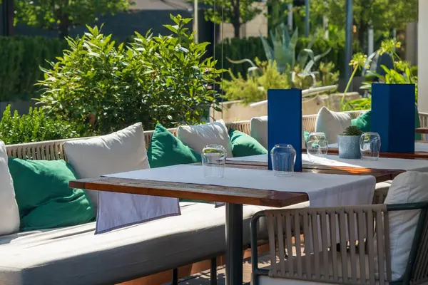 Yastıklı banklar, yeşil ve beyaz yastıklar ve yemyeşil güneş ışığı altında yemyeşil masalarla çevrili rahat bir açık hava restoranı..