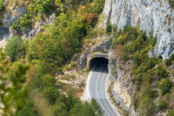 Yoğun orman ve engebeli araziyle çevrili sarp kayalıkların arasından oyulmuş bir tünele giden kavisli bir yol..
