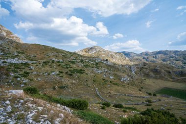 Kayalık yamaçları ve çimenli vadileri olan engebeli bir dağ manzarası. Dağılmış bulutlarla parlak mavi bir gökyüzüne karşı kurulmuş. Huzurlu, manzaralı bir manzara yaratıyor..