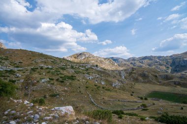 Dağılmış bulutlarla dolu parlak mavi bir gökyüzünün altında kayalık çıkıntıları ve çimenli arazisi olan engebeli bir dağ silsilesi dramatik bir manzara yaratıyor..