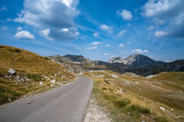 Manzaralı, dolambaçlı bir yol engebeli bir dağ manzarasını kesiyor. Parlak mavi bir gökyüzünün altında, dağınık bulutlarla..