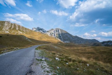 Dolambaçlı kırsal bir yol çimenli tepelerden geçerek dağlık tepelere, parlak mavi bir gökyüzünün altında dağınık bulutlarla ilerliyor..