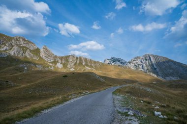 Dolambaçlı bir yol, çimenli tepelerden ve pürüzlü kayalıklardan geçiyor. Parlak mavi bir gökyüzünün altında, dağınık bulutlarla..