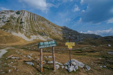 Trail signs in Durmitor National Park stand against a backdrop of rugged rocky mountains and grassy terrain under a blue sky. clipart