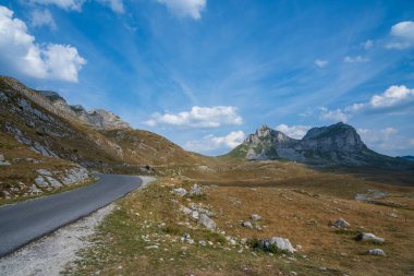 Dolambaçlı bir yol engebeli bir dağ manzarasını keser kayalık tepeler ve sarp uçurumlar dağınık mavi bulutlarla dolu bir gökyüzünün altında.