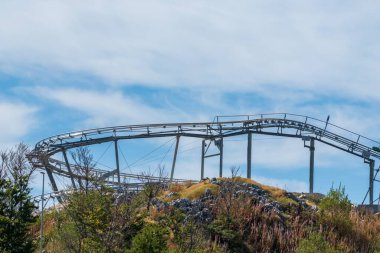 Boş bir roller coaster pisti kayalık bir dağ yamacının üzerinde kıvrımlı kıvrımlı, parlak mavi bir gökyüzüne karşı ince bulutlu.