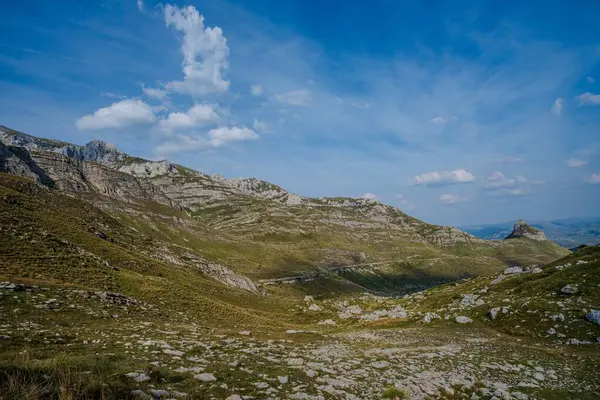 Uçsuz bucaksız bir dağ vadisi kayalık yamaçlar ve yeşil yamalarla, berrak mavi bir gökyüzünün altında dağınık bulutlarla, sakin vahşi doğayı gözler önüne seriyor..