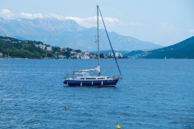 Sakin kıyı sularına demir atmış tek bir yelkenli uzak dağları ve arka planda görünen bir kıyı kasabası var..