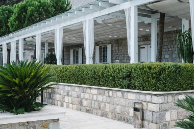 Gölgeli pergola ve beyaz perdeleri olan, yemyeşil bitkilerle çevrili taştan bir bina ve düzgün kesilmiş bir çit..