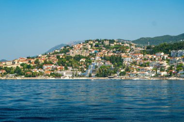 Kotor Körfezi, Karadağ - 22 Ağustos 2024: Güneşli bir günde, kıyı şeridinde kırmızı kiremitli çatıları, sahile bakan bir güvertesi ve sakin mavi suları olan manzaralı bir kasaba.