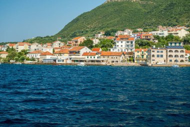 Kotor Körfezi, Karadağ - 22 Ağustos 2024: Deniz kenarında evleri, küçük plajı ve güneşli bir gökyüzünün altında açık mavi denizi olan büyüleyici kıyı köyü.