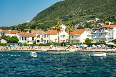Kotor Körfezi, Karadağ - 22 Ağustos 2024: Durgun bir kıyı şeridi boyunca şemsiyeli ve güneşli deniz evleri, açık bir gökyüzü altında yüzücüler ve demirlemiş teknelerin yer aldığı.