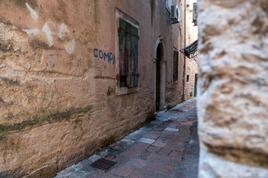 Kotor Körfezi, Karadağ - 22 Ağustos 2024: Tarihi bir Akdeniz kasabasında yıpranmış taş duvarlar, graffitiler ve kapalı ahşap panjurlarla dolu dar bir sokak.