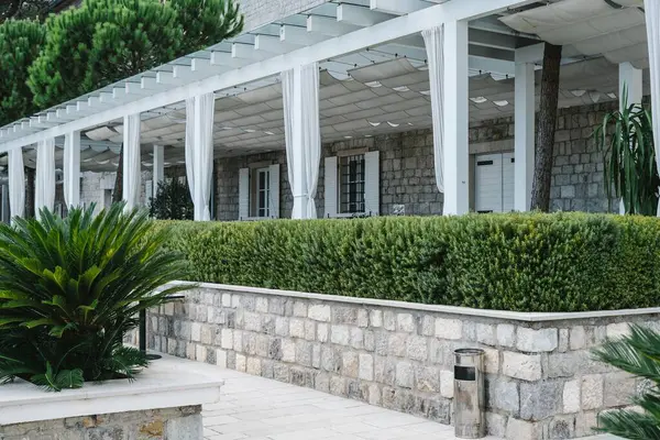 Gölgeli pergola ve beyaz perdeleri olan, yemyeşil bitkilerle çevrili taştan bir bina ve düzgün kesilmiş bir çit..