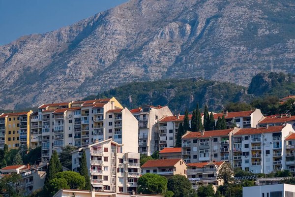 Bay of Kotor, Montenegro - August 22, 2024: A cluster of apartment buildings with red-tiled roofs nestled at the base of a rugged mountain, surrounded by greenery in a Mediterranean town.