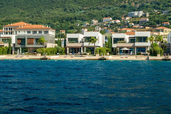 Залив Котор, Монтенегро - 22 августа 2024 года: роскошные фронтальные виллы с современной архитектурой, доступом к частному пляжу и потрясающим видом на море, на фоне пышной гильотины.
