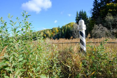 Close-up of a black-and-white border post surrounded by tall autumn vegetation and a forested backdrop under a blue sky. clipart