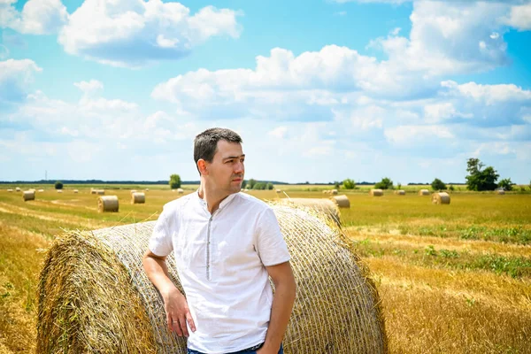 Happy farmer is standing beside bales of hay. He is examining straw after successful harvesting. Strong handsome man portrait on agricultural field.