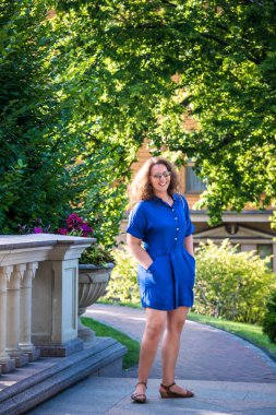 Şehir parkında güneş gözlüklü genç ve güzel bir kadın. Yaz günü elbise giyip kameraya bakıyor..