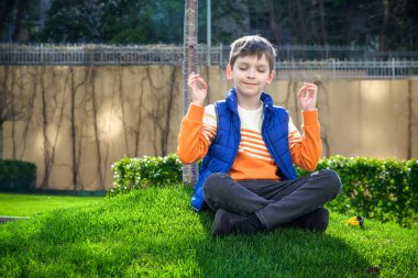 Taze yeşil çimen tarlasında oturan beyaz çocuk günlük giysiler içinde meditasyon yapıyor, mutlu çocuk dışarıda huzurlu bir yaz haftasonunun tadını çıkarıyor. Rahatlama ilham konsepti.