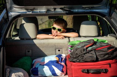 Arabanın bagajında oturan güneş gözlüklü sevimli çocuk. Mutlu çocuk portresi açık araba bagajı ile ebeveynin mesleğine hazırlanmasını bekliyor. Aile gezisi araba konseptiyle yapılır..