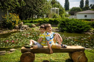 İki erkek kardeş, barışçıl göl kenarındaki parkta bir bankta Lily Nymphaeum 'la oturuyorlar. Çocuklar sıcak yaz gününde doğada dinleniyorlar..