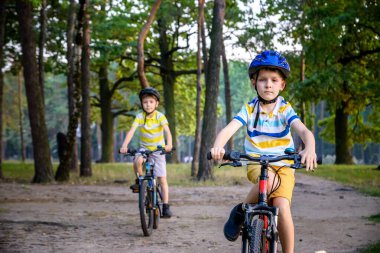 İki küçük çocuk yaz parkında bisiklet sürerken günlük kıyafetlerini giymiş. Doğada güneşli sonbahar gününde bisiklet süren aktif çocuklar. Güvenlik, spor, çocuk konseptiyle eğlence..