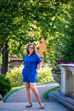 Şehir parkında güneş gözlüklü genç ve güzel bir kadın. Yaz günü elbise giyip kameraya bakıyor..