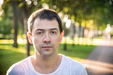 Açık havada, beyaz tişörtlü, doğa parkının arka planında güzel, ciddi bir adamın portresini çek. Moda modeli.