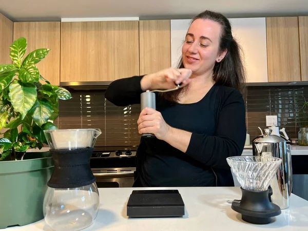 Stock image A girl in her hands with a manual grinder grinds coffee beans There is coffee tree near it and on the white countertop in the kitchen there are various devices for a craft drink Relax coffee house ad